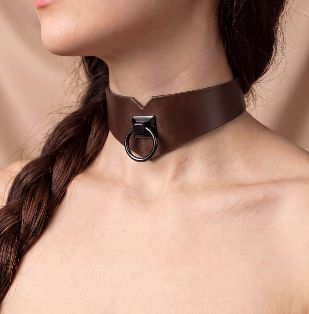 A close-up of a woman's neck and chest area wearing a brown leather choker necklace with a metal ring.