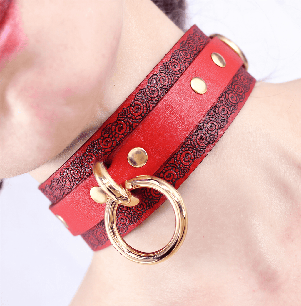 A close-up of a red leather choker necklace with gold-colored metal rings and a decorative pattern on the leather.