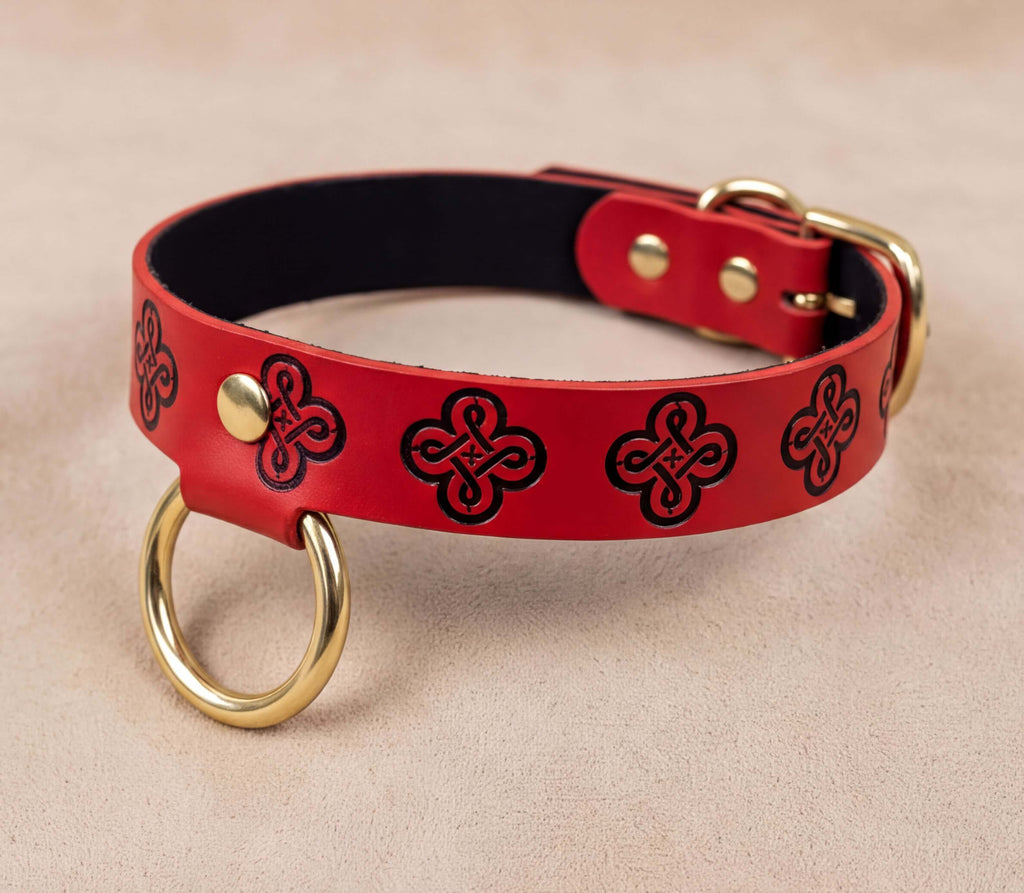A red leather collar with a black and gold design, featuring a gold ring and buckle, is shown on a beige surface.