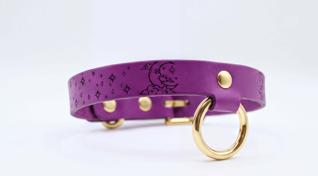 A purple leather collar with a moon and star design, featuring a gold-colored metal ring and buckle.