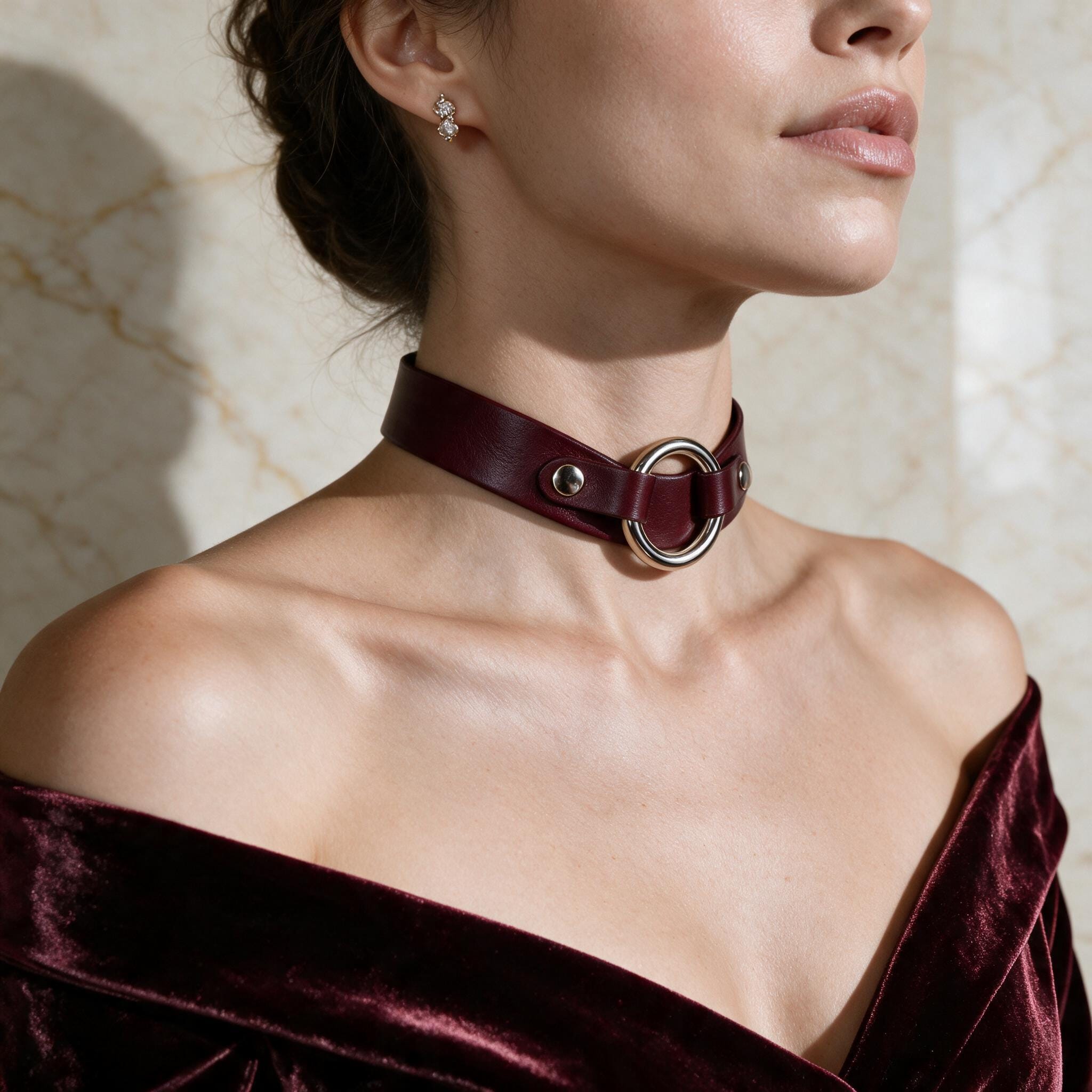 A woman wearing a burgundy choker necklace with a large circular buckle, posing against a marble wall.