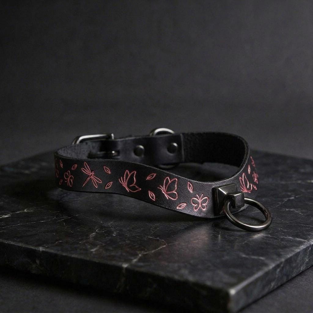 A black leather collar with pink floral designs and a metal ring on the front, resting on a dark surface.
