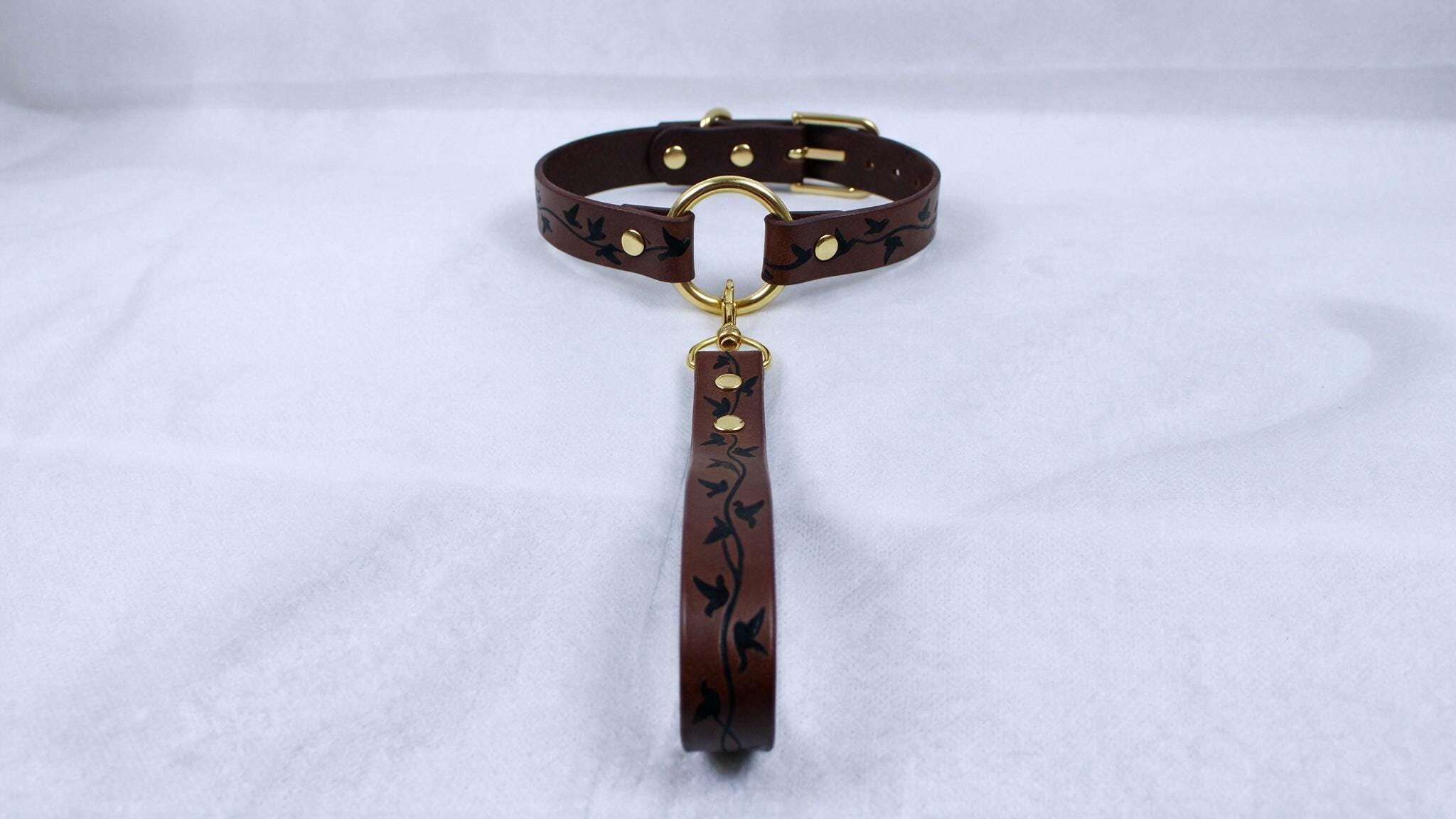 A brown leather collar with a gold-colored buckle and a black floral pattern on the strap.