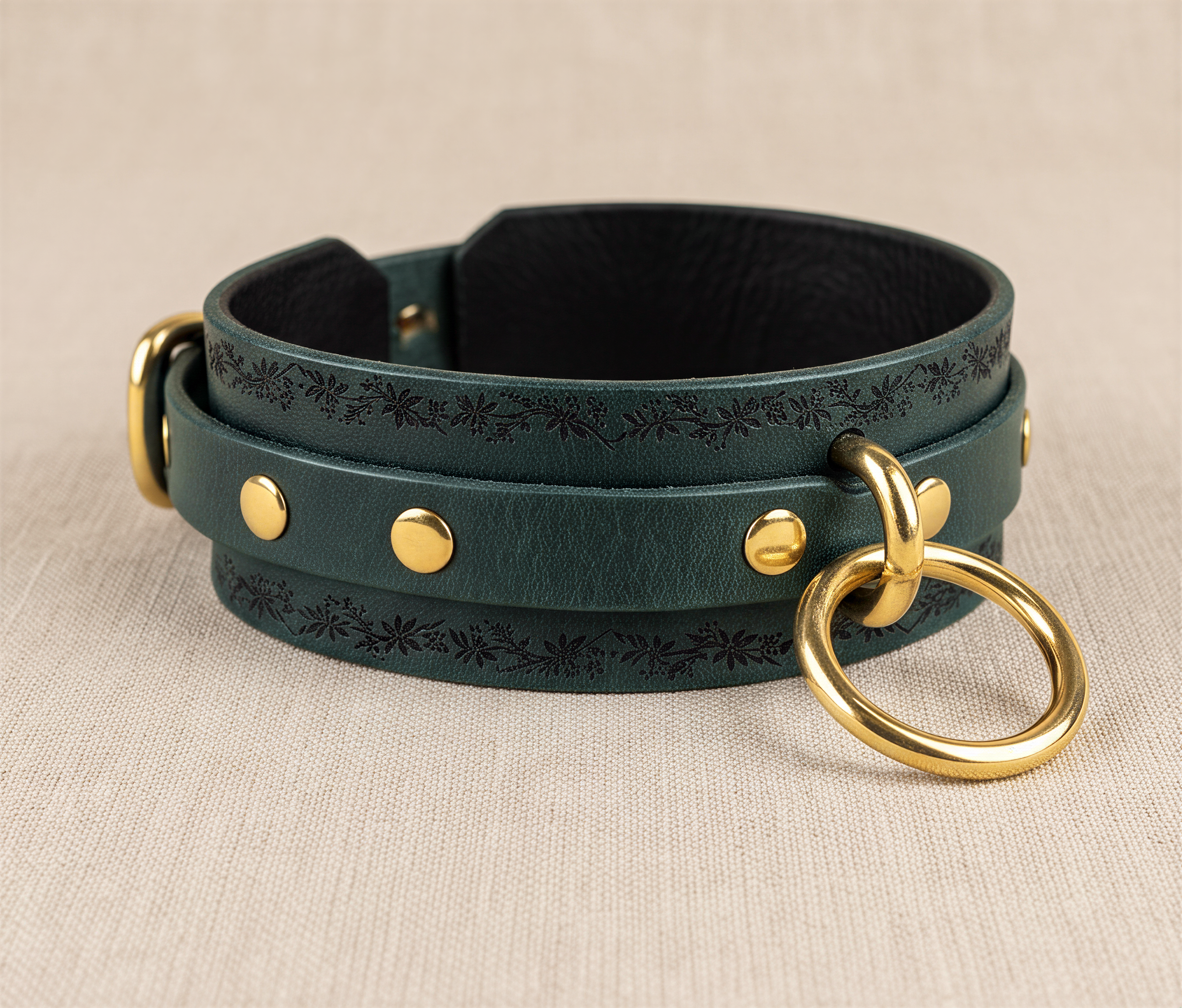 A dark green leather choker collar with gold-colored metal rings and studs, resting on a beige background.