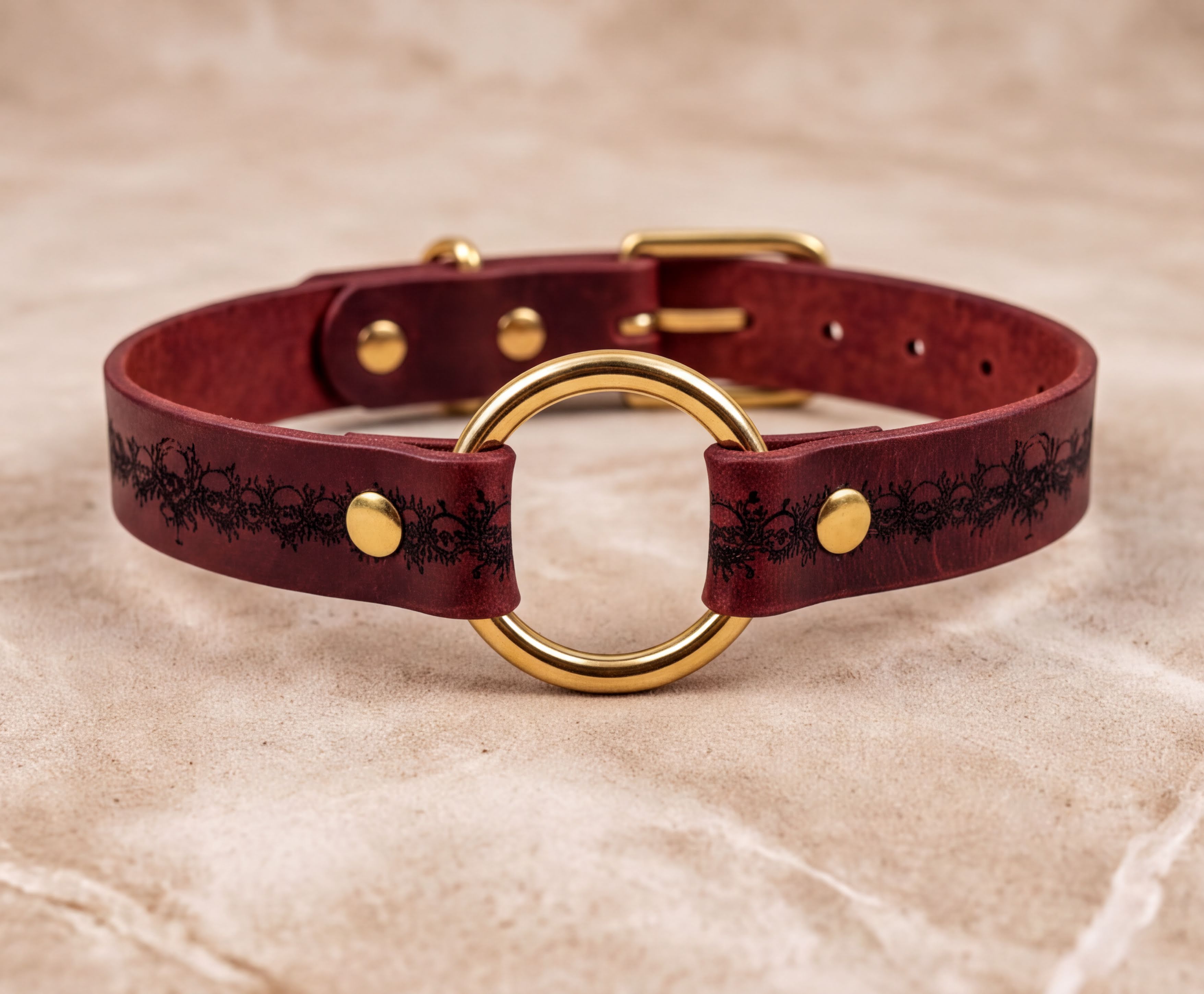 A burgundy leather collar with a gold-colored circular buckle and a decorative pattern on the leather.