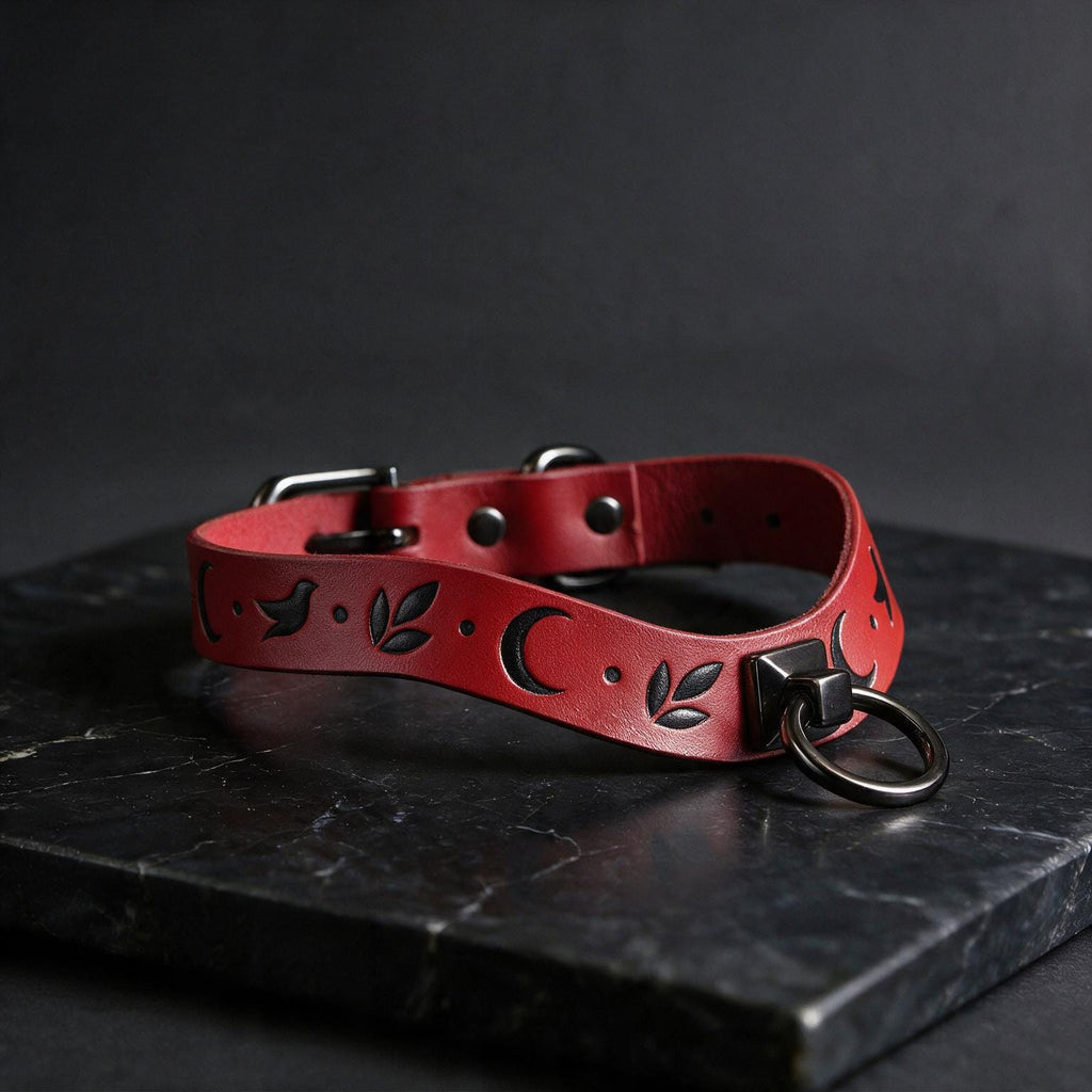 A red leather collar with a crescent moon and leaf design, featuring a metal ring and buckle, is displayed on a black marble surface.