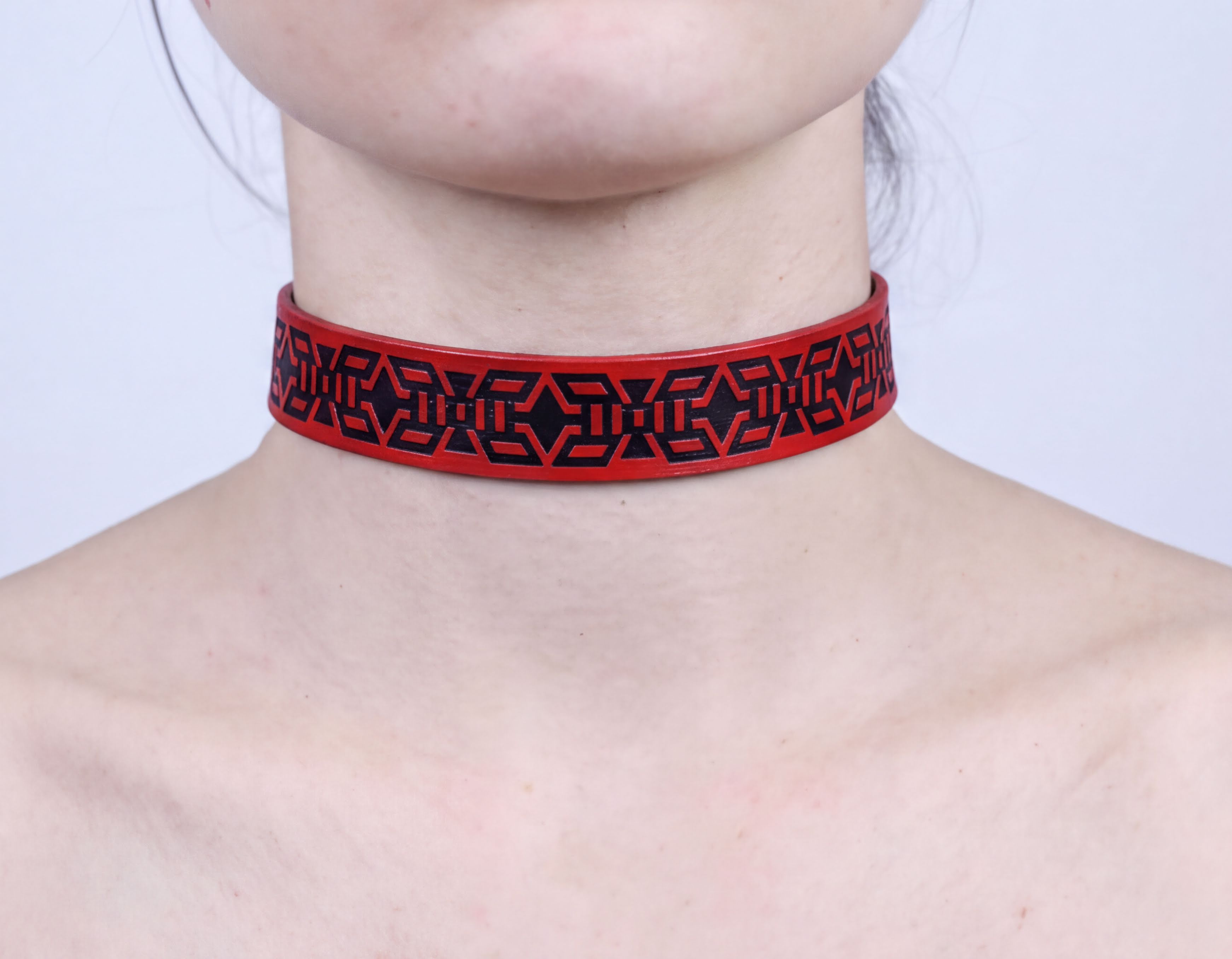 A close-up of a person's neck wearing a red choker necklace with a black geometric pattern.
