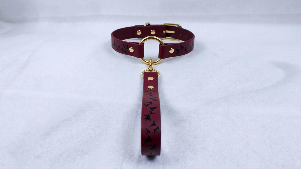 A burgundy leather collar with a gold-colored buckle and a black bird design on the strap is displayed on a white surface.
