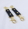 A pair of black leather straps with gold metal loops and hooks, arranged side by side on a white background.