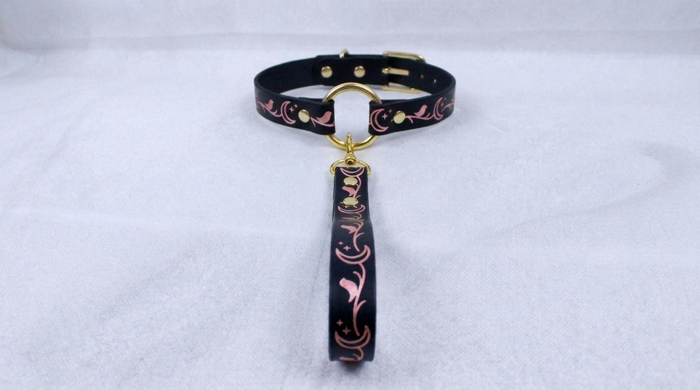 A black collar with a gold-colored buckle and a pink floral pattern on the strap is displayed against a white background.