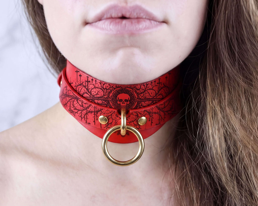 A close-up of a person's neck and chin wearing a red choker necklace with a skull design and a gold ring.