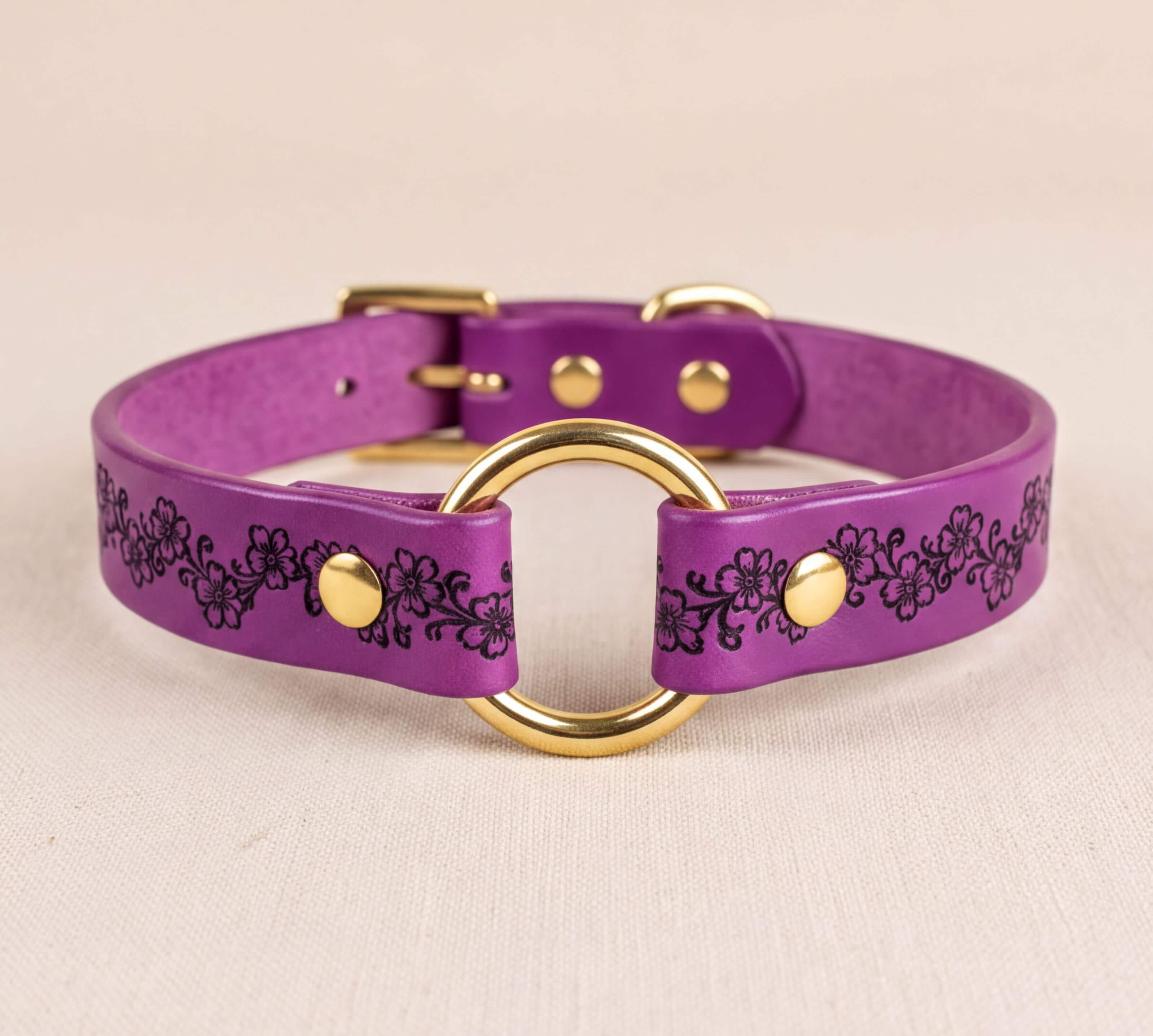 A purple leather collar with a gold-colored metal ring and decorative floral patterns.