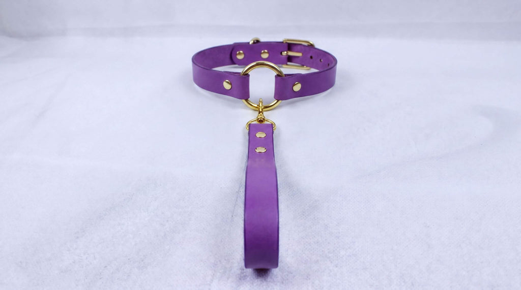 A purple leather collar with a gold-colored buckle and a long strap is displayed on a white surface.