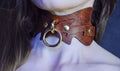 A close-up of a person's neck wearing a brown leather choker necklace with a metal ring and two gold-colored buttons.