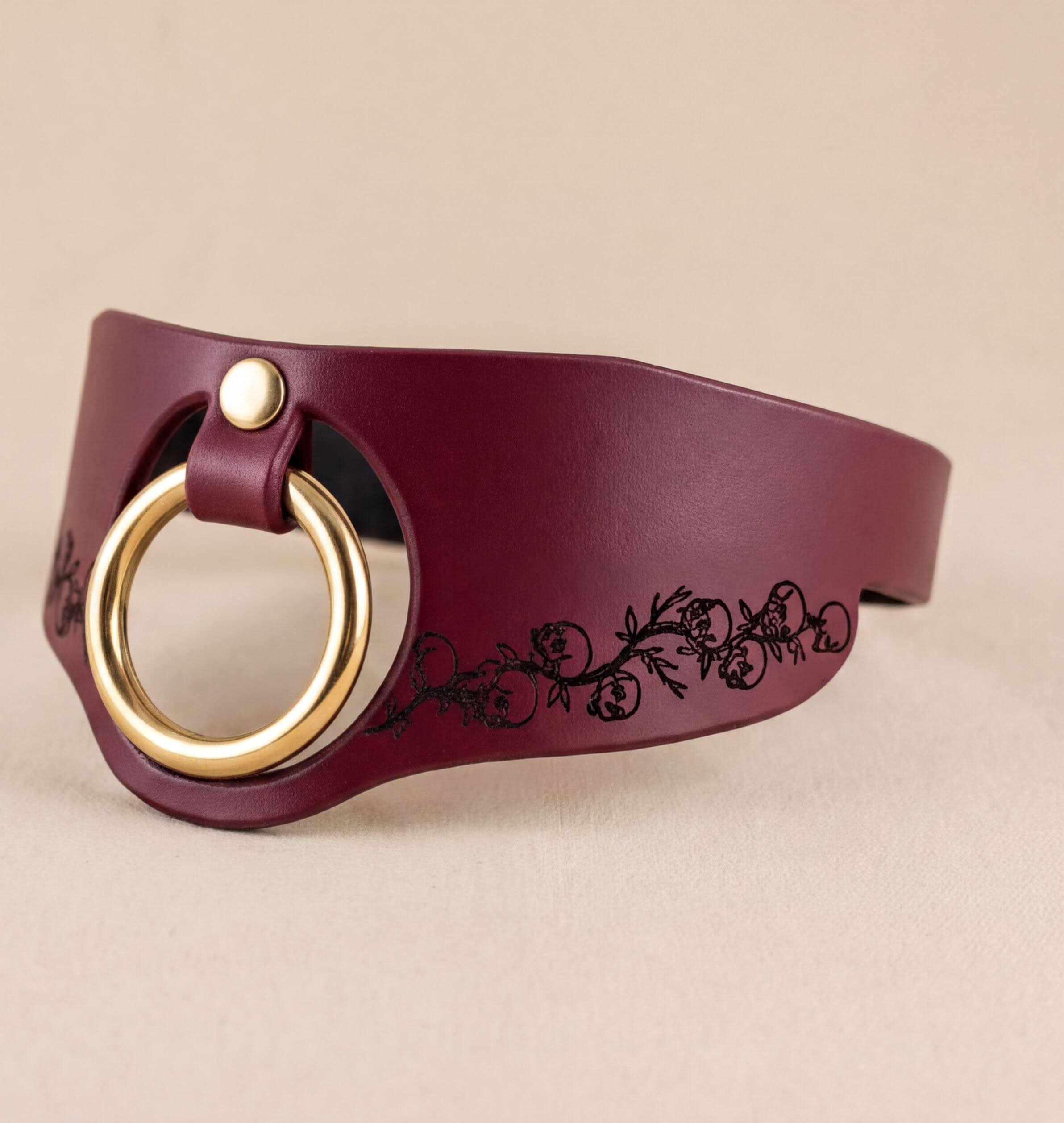 A burgundy leather choker necklace with a gold-colored ring and a floral design on the side.