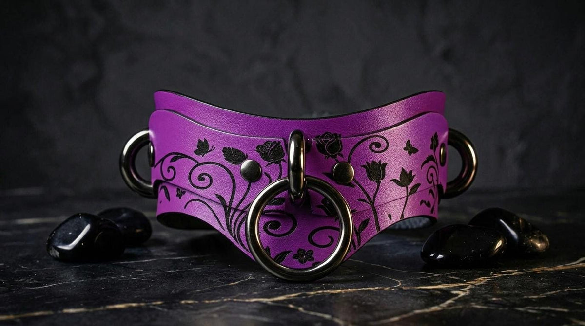 A purple leather collar with black floral designs and metal rings is displayed on a dark surface, accompanied by black crystals.