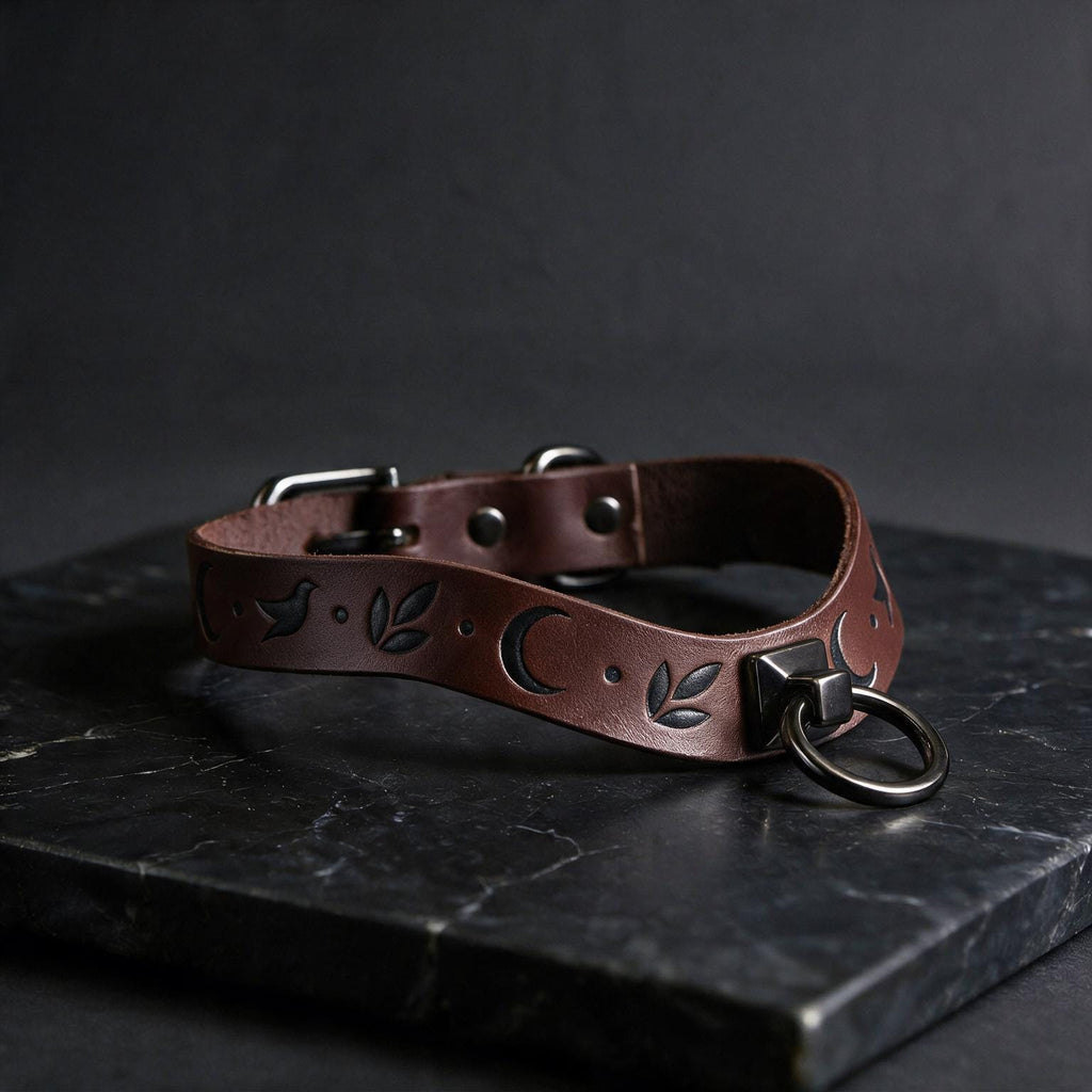 A brown leather collar with a moon and leaf design, featuring a metal ring and buckle, is displayed on a black marble surface.