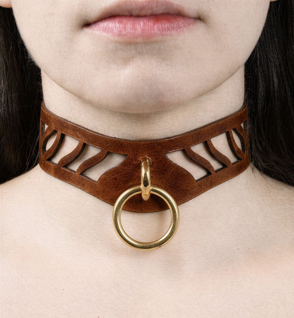 A close-up of a person's neck and chest area, featuring a brown leather choker necklace with a gold ring and a cutout design.