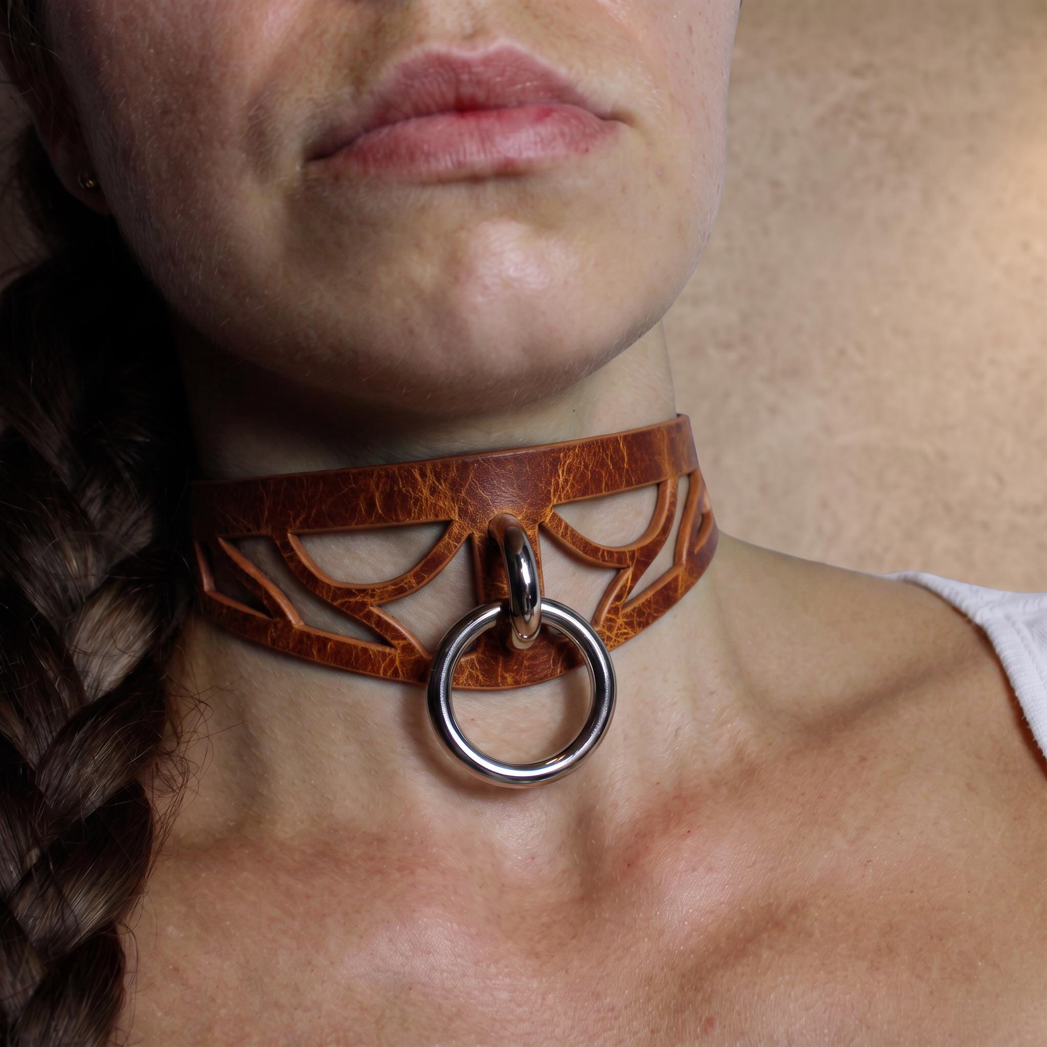 A close-up of a woman's neck and chest area, featuring a leather choker necklace with a metal ring and a braided hairstyle.