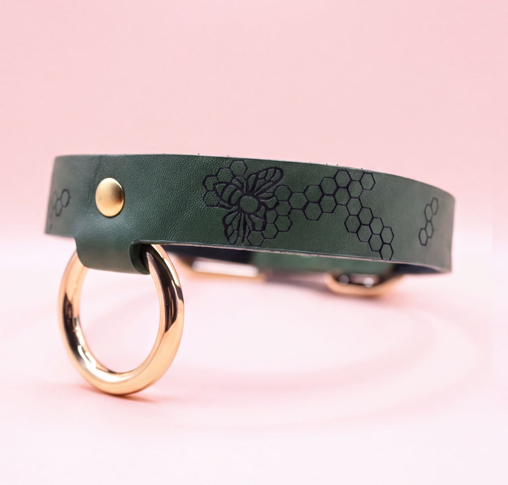 A green leather collar with a gold ring and a bee design on it, set against a pink background.