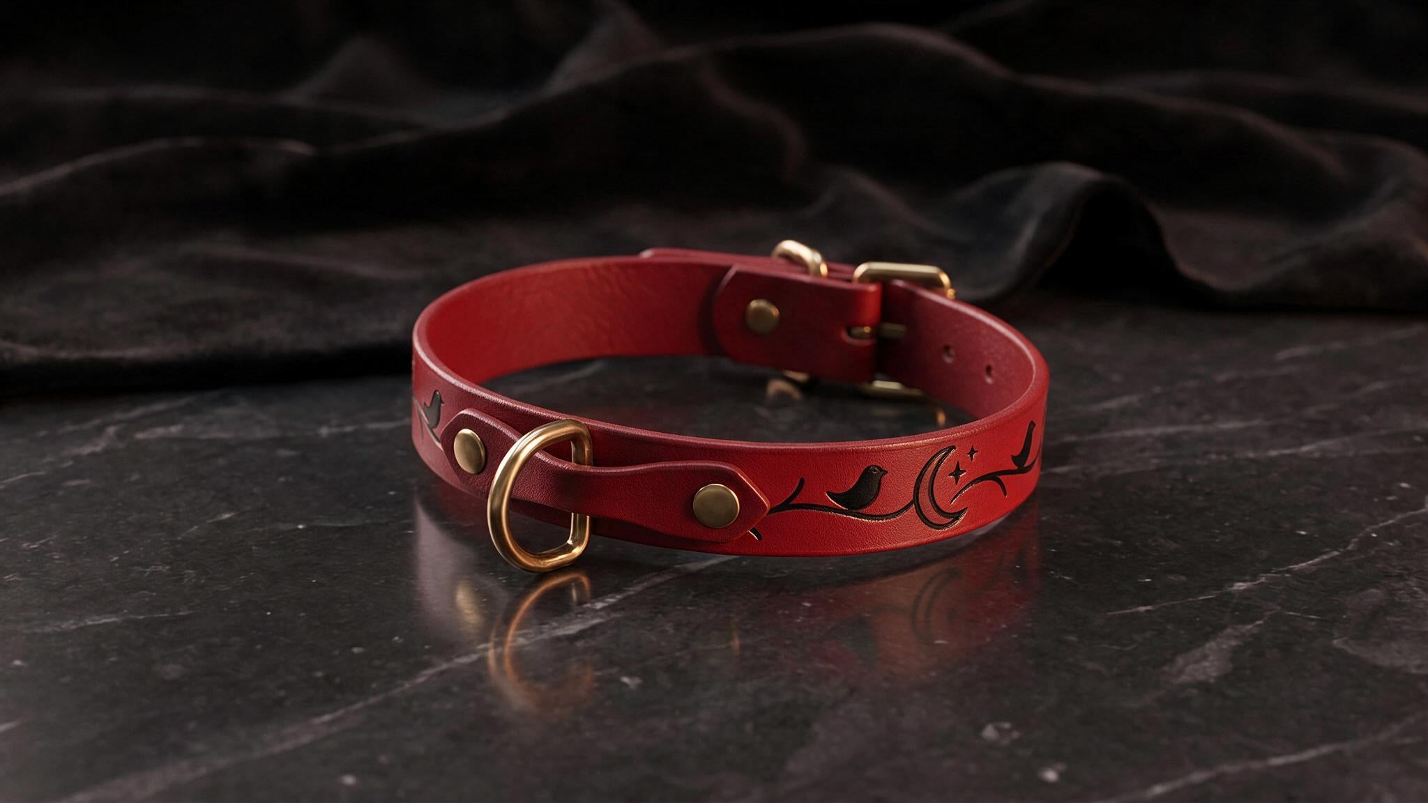 A red leather collar with a crescent moon and bird design, featuring a gold-colored buckle, is displayed on a dark surface.