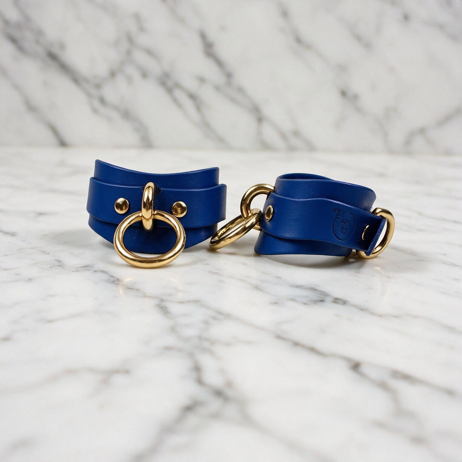 A pair of blue leather wrist straps with gold metal rings and buckles, placed on a marble surface.