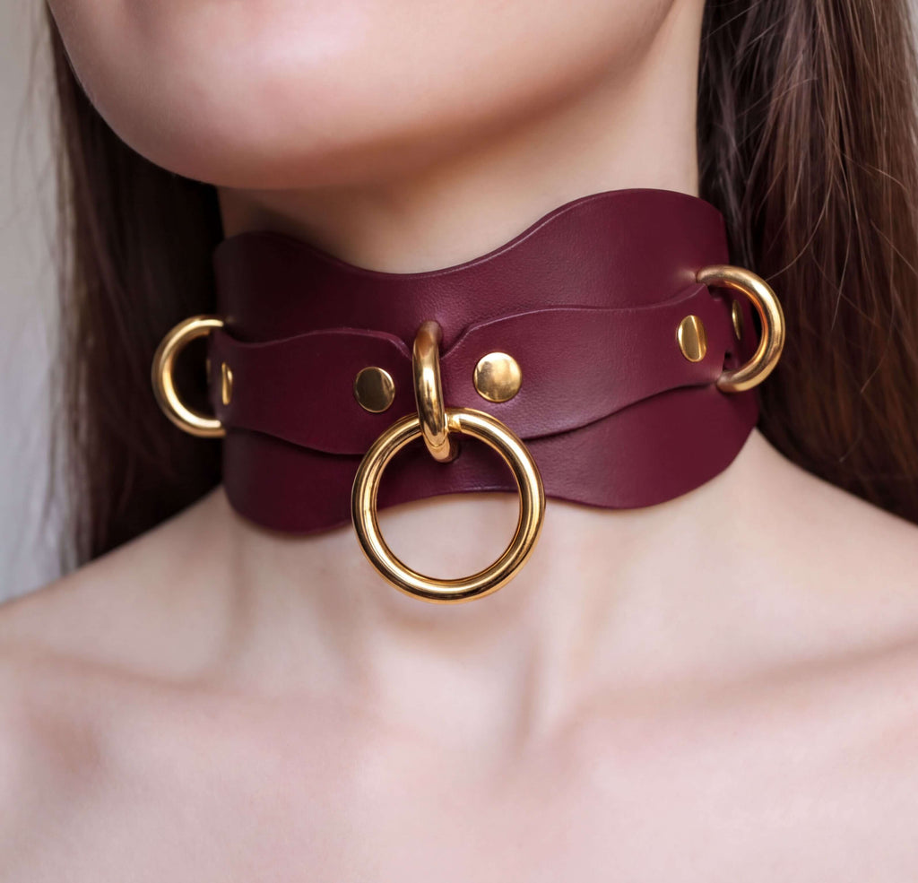 A close-up of a person's neck wearing a burgundy leather choker necklace with gold-colored metal rings.