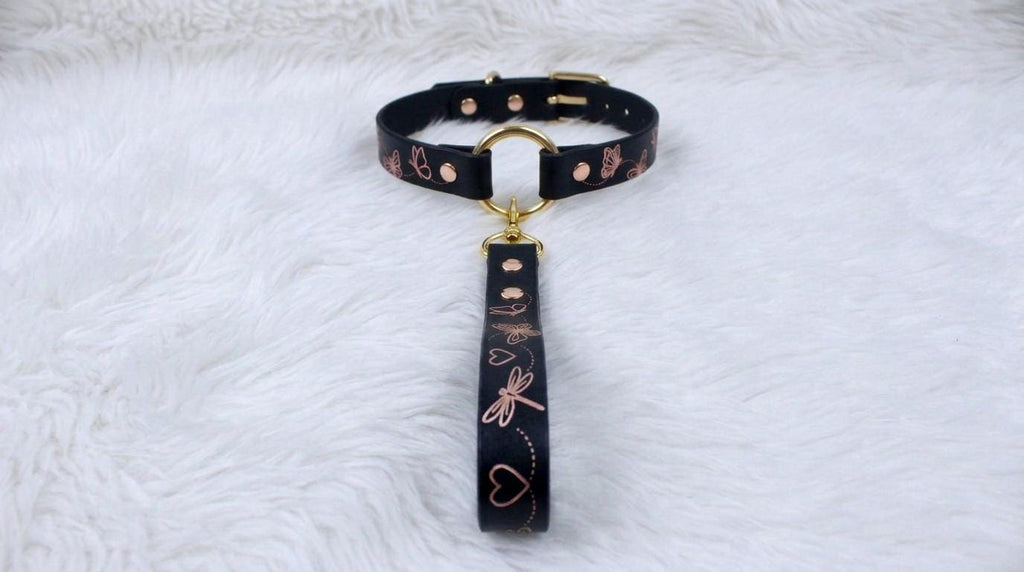 A black collar with a gold-colored buckle and a handle attached to it, placed on a white furry surface.