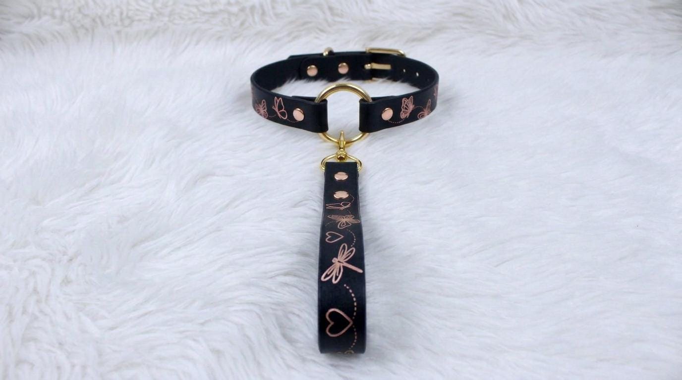 A black collar with a gold-colored buckle and a handle attached to it, placed on a white furry surface.