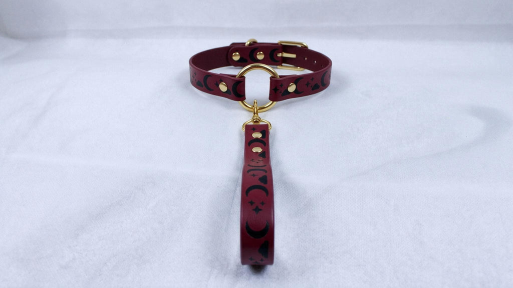 A burgundy leather collar with a gold-colored buckle and a dangling handle is displayed on a white surface.