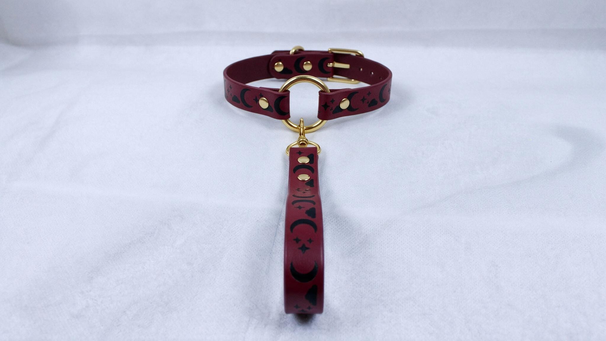 A burgundy leather collar with a gold-colored buckle and a dangling handle is displayed on a white surface.
