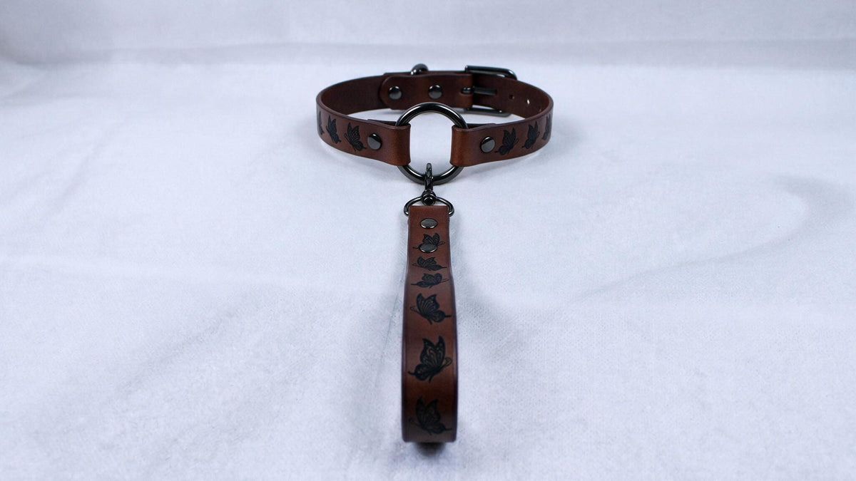 A brown leather collar with a metal buckle and a handle attached to it, resting on a white surface.