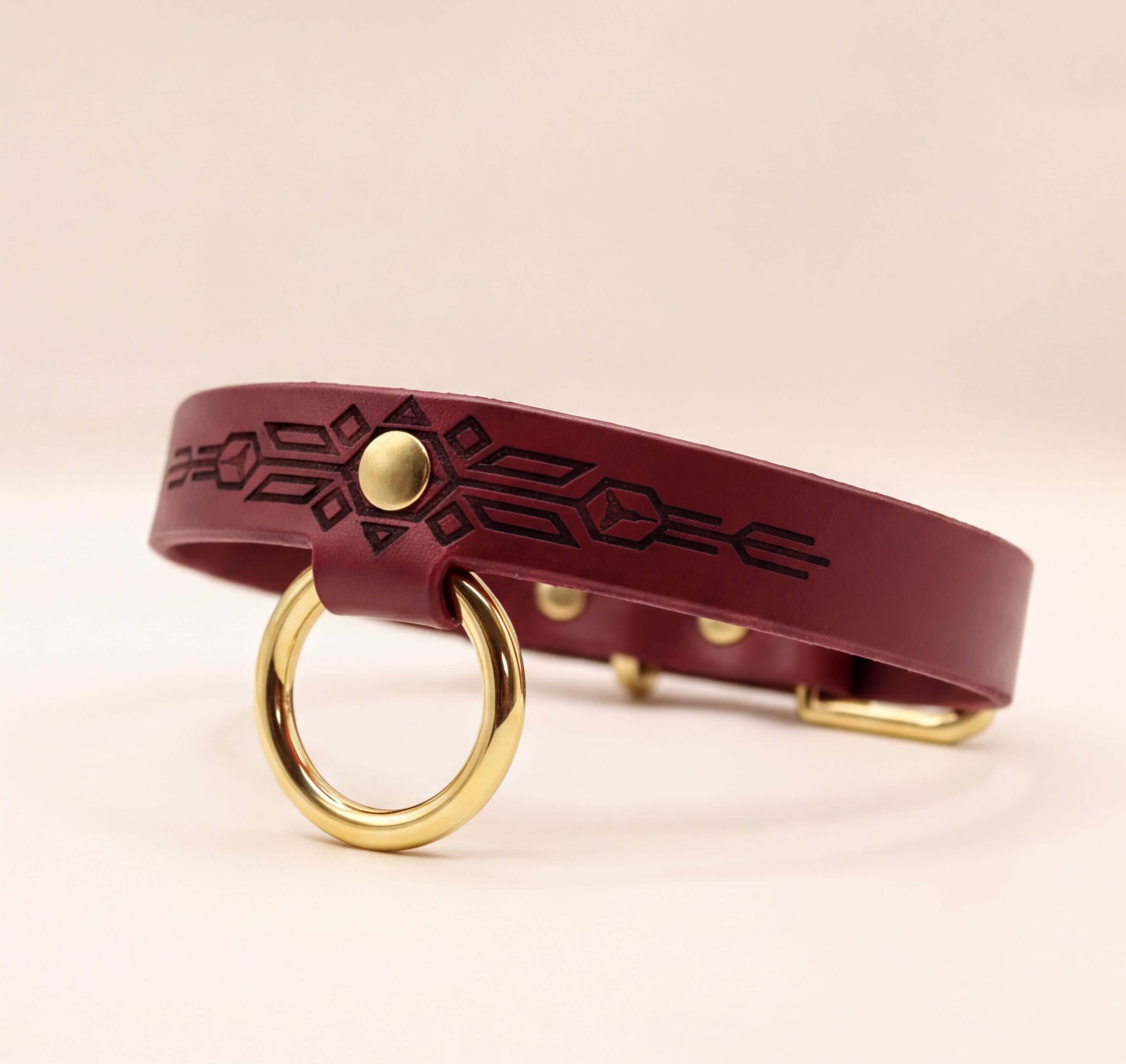 A burgundy leather collar with a gold-colored metal ring and a geometric pattern on the strap.