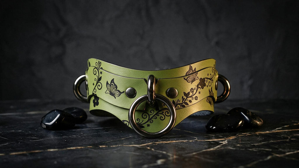 A green leather choker necklace with black floral and butterfly designs, featuring two metal rings and black stones on a dark surface.