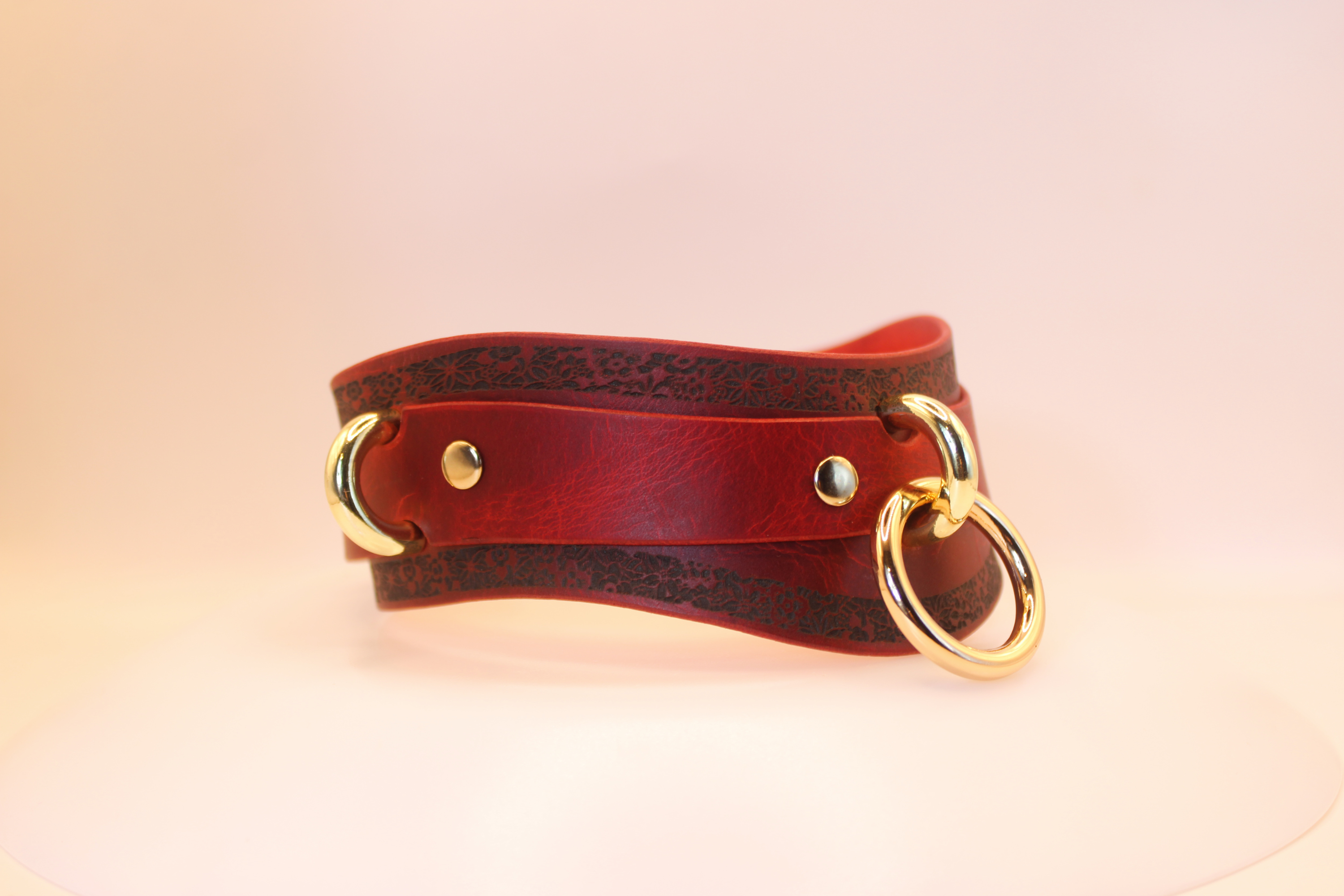 A red leather choker necklace with gold-colored metal rings and a decorative pattern on the surface.