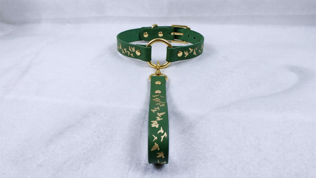 A green leather collar with a gold-colored buckle and a gold-colored tag attached to it, resting on a white surface.