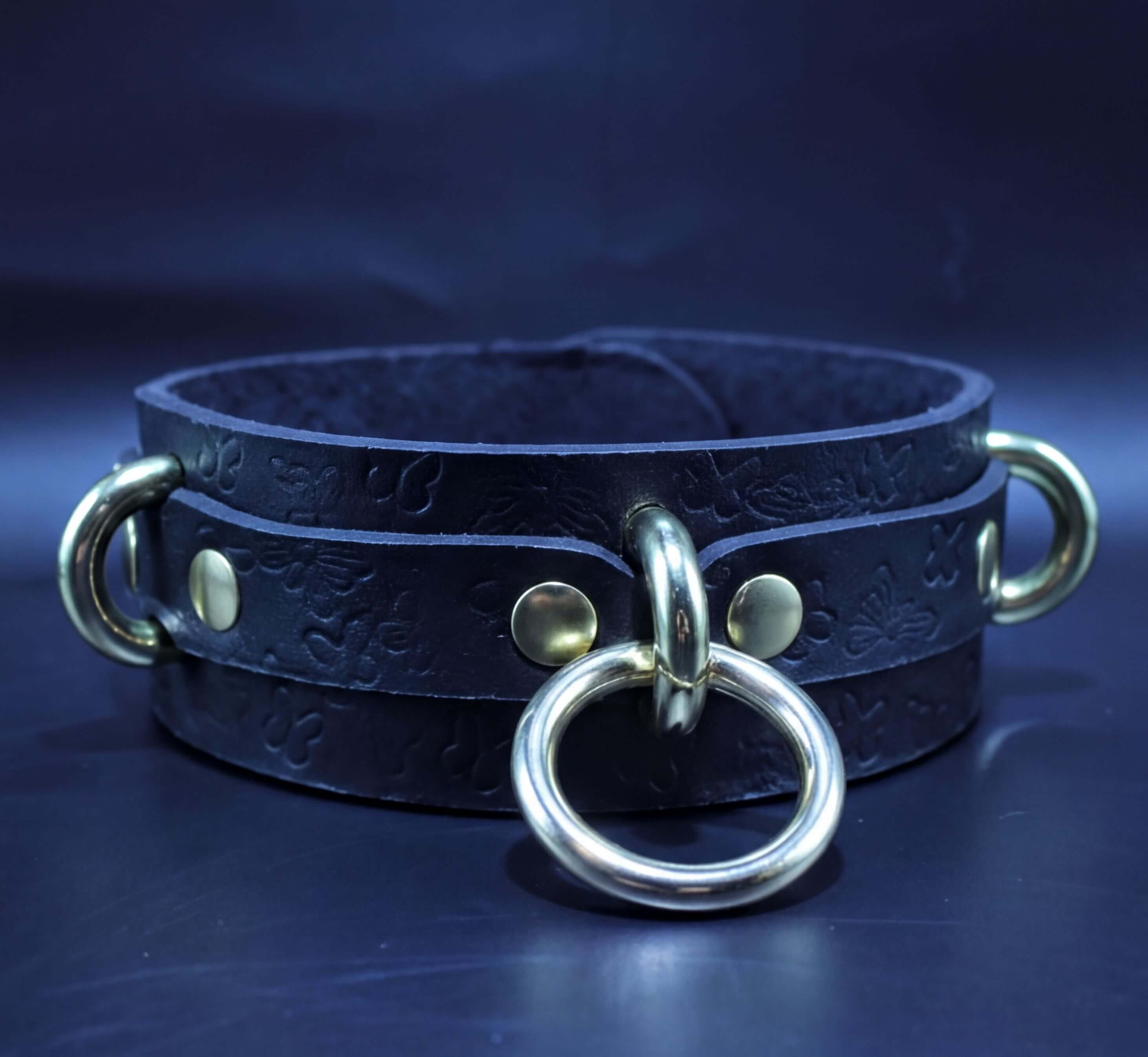 A black leather choker necklace with a metal ring and two metal rings on either side, set against a dark blue background.