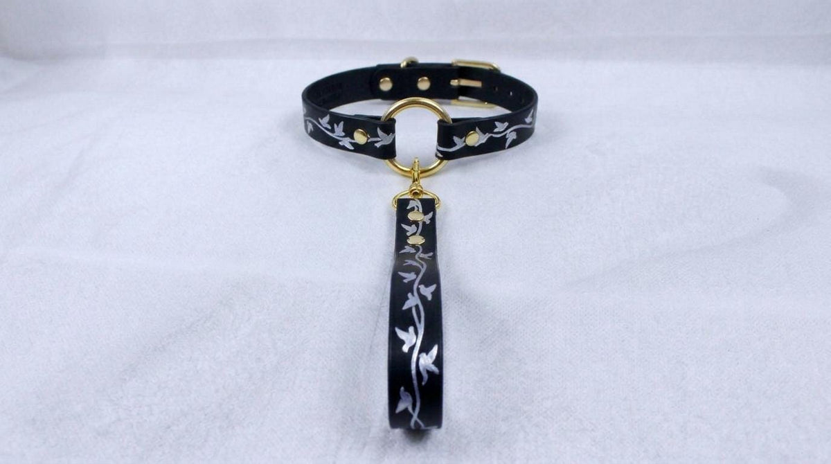 A black collar with a gold-colored buckle and a white floral pattern on the strap is displayed against a white background.