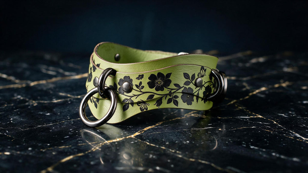 A green leather choker necklace with black floral designs and metal rings is displayed on a black marble surface.