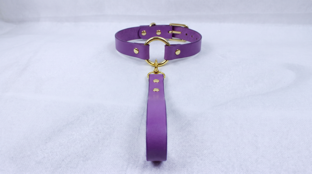 A purple leather collar with a gold-colored buckle and a long strap is displayed on a white surface.