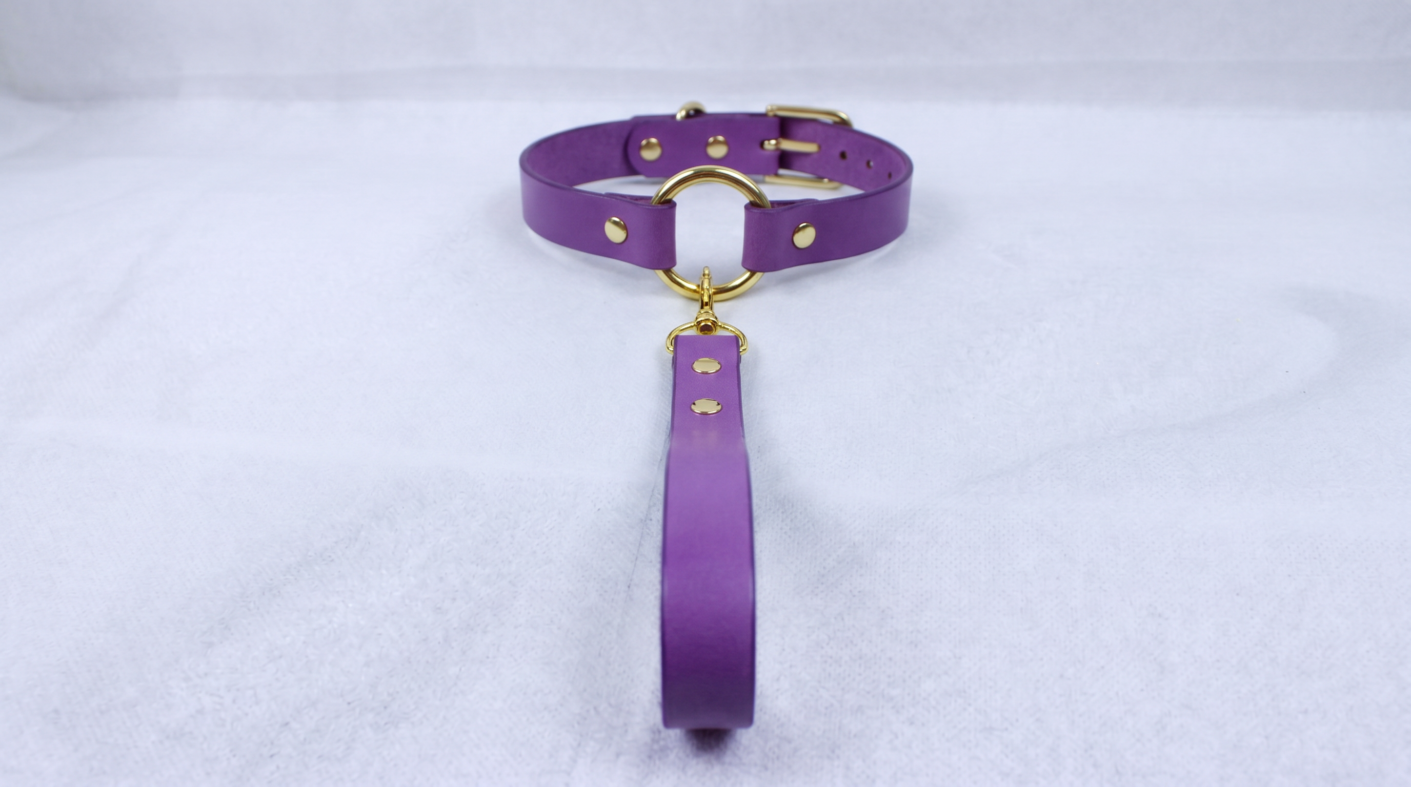 A purple leather collar with a gold-colored buckle and a long strap is displayed on a white surface.