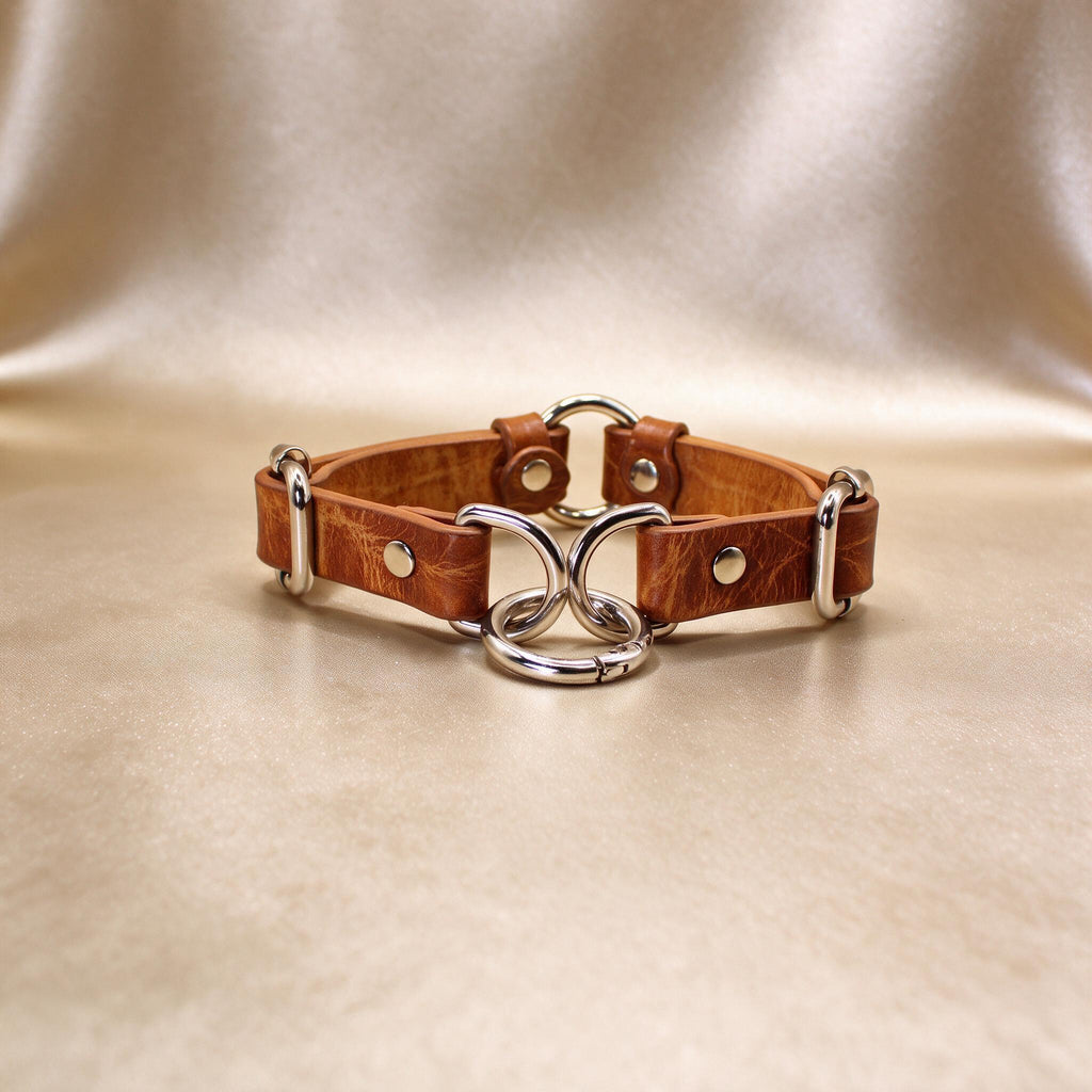 A brown leather collar with metal rings and buckles, displayed on a beige background.