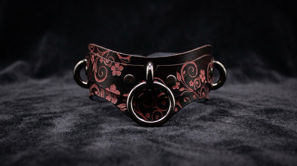 A dark leather choker necklace with a floral pattern and metal rings on the front.