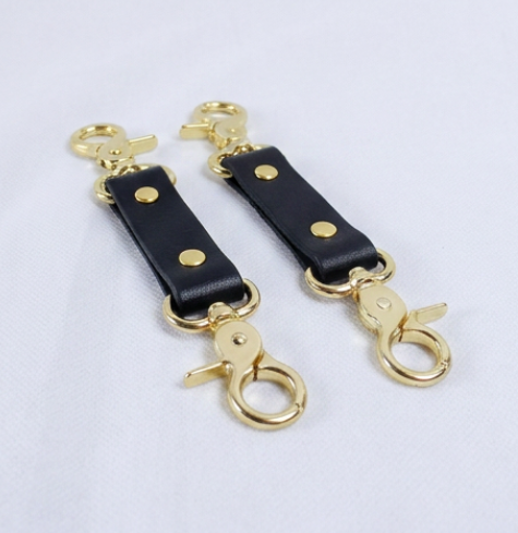 A pair of black leather straps with gold metal loops and hooks, arranged side by side on a white background.