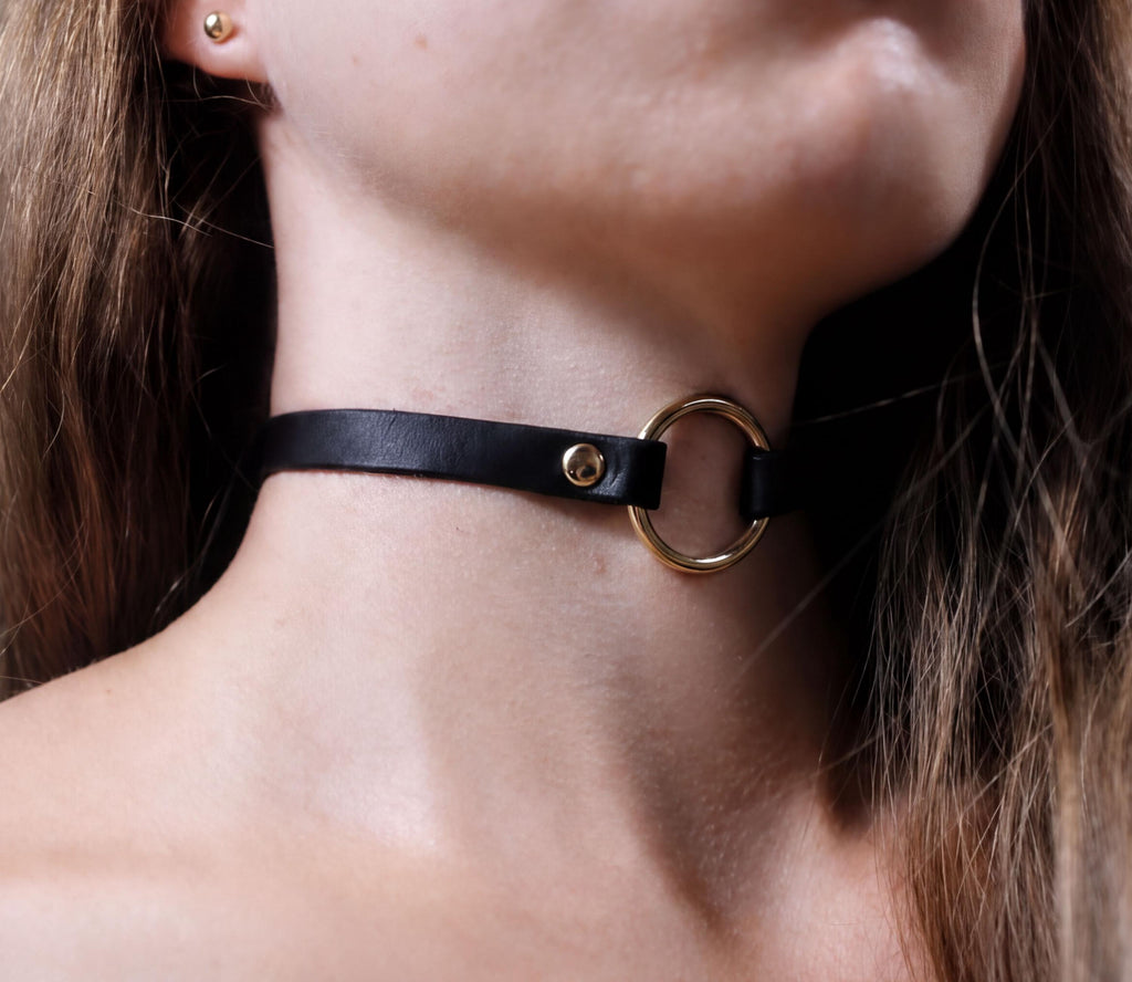 A close-up of a person's neck and chin wearing a black leather choker necklace with a gold ring.