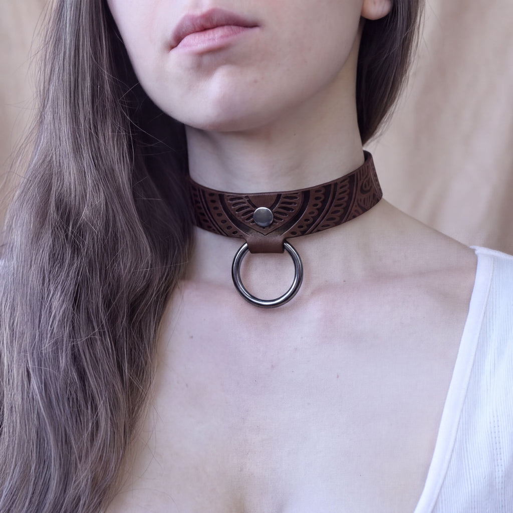 A close-up of a woman's neck and chest area, featuring a leather choker necklace with a metal ring.