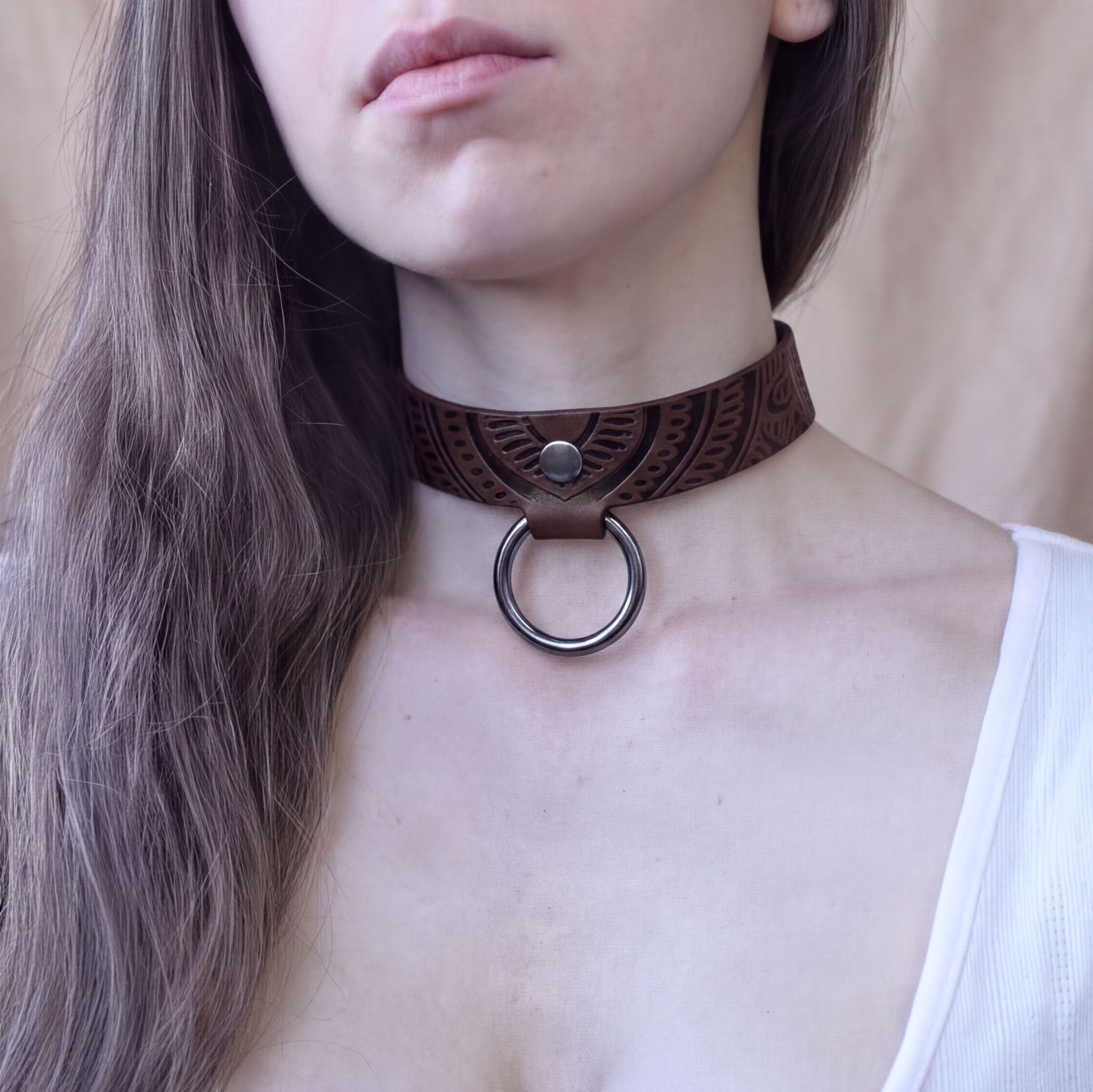 A close-up of a woman's neck and chest area, featuring a leather choker necklace with a metal ring.