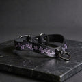 A black leather collar with a purple leaf design on it, resting on a dark surface.