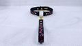A black leather collar with a gold-colored buckle and a pink floral pattern on the strap is displayed on a white surface.