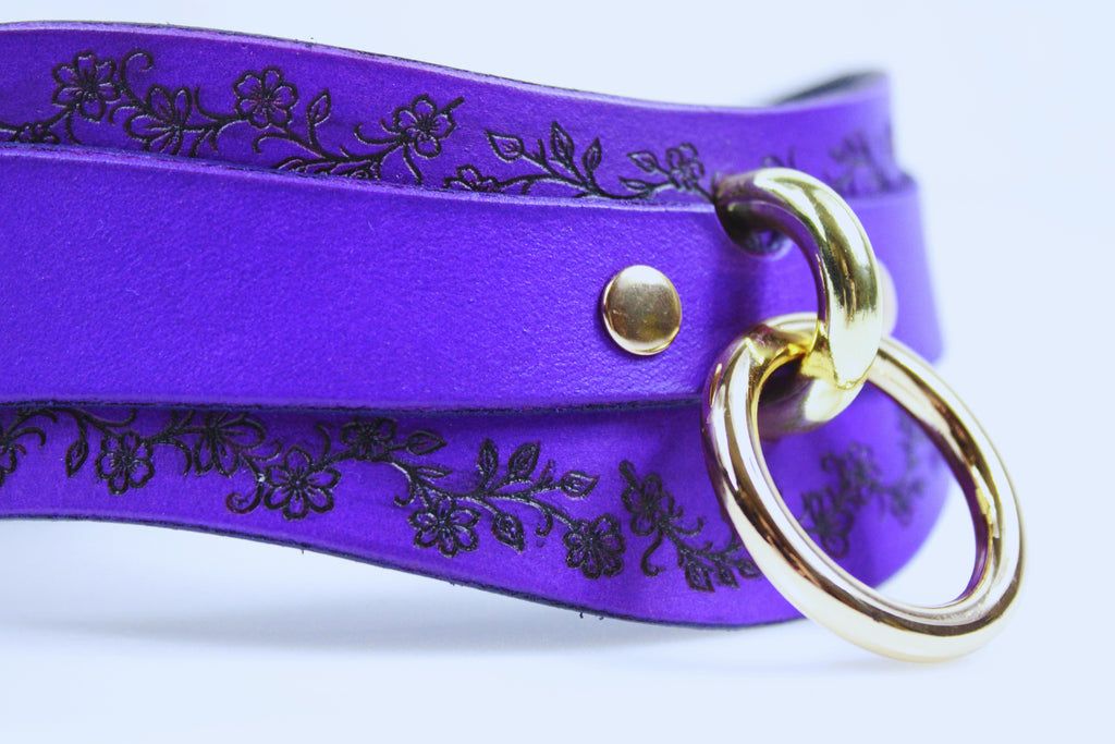 A close-up view of a purple leather choker necklace with a gold-colored ring and a floral design on the clasp.